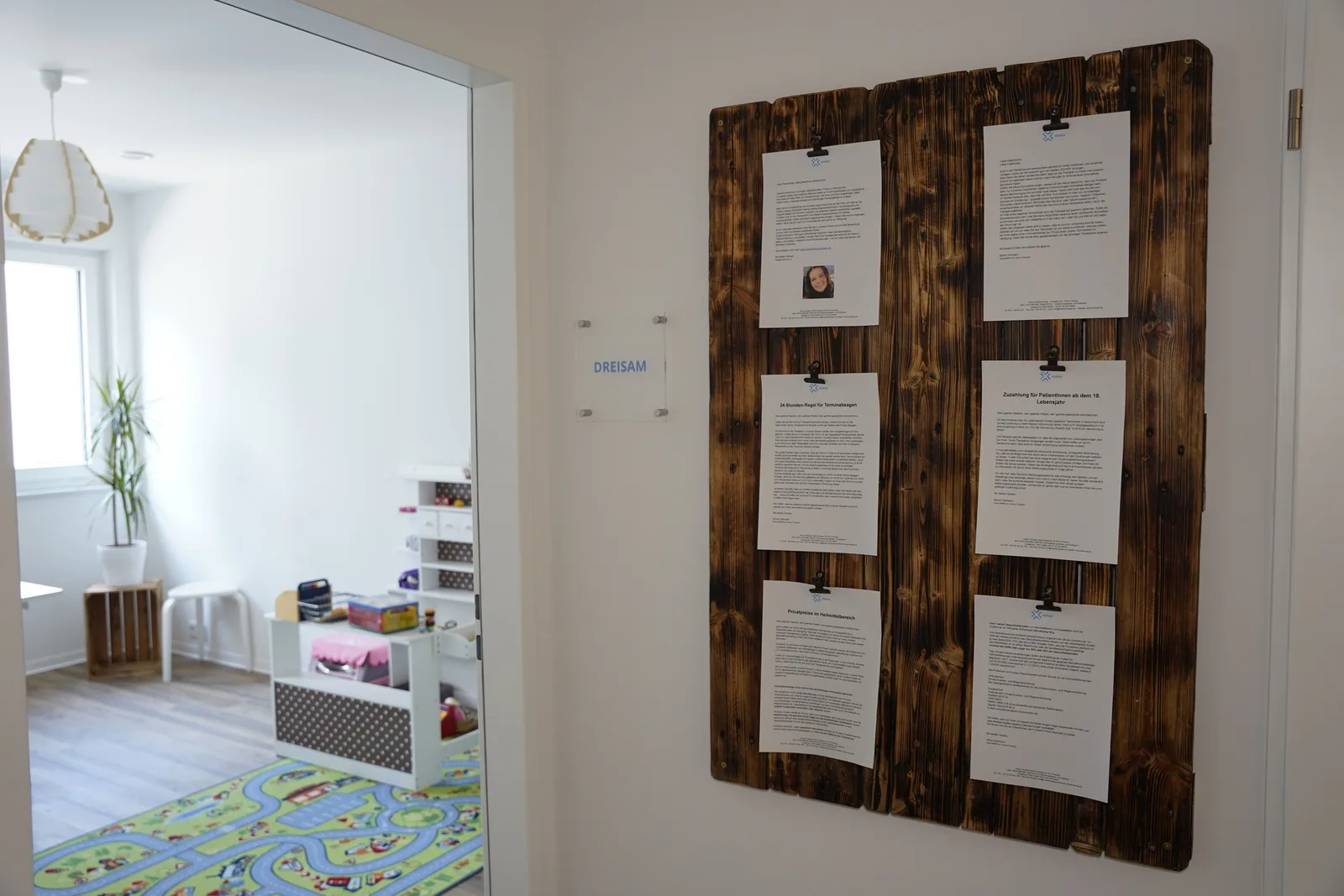 Praxisflur mit einer Holztafel voller Aushänge neben einer offenen Therapieraumtür